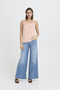 Ženska, oblečena v svetlo rožnato camisolo z čipkastim robom, široke modre kavbojke in rjave Mary Jane čevlje, stoji na preprostem ozadju.