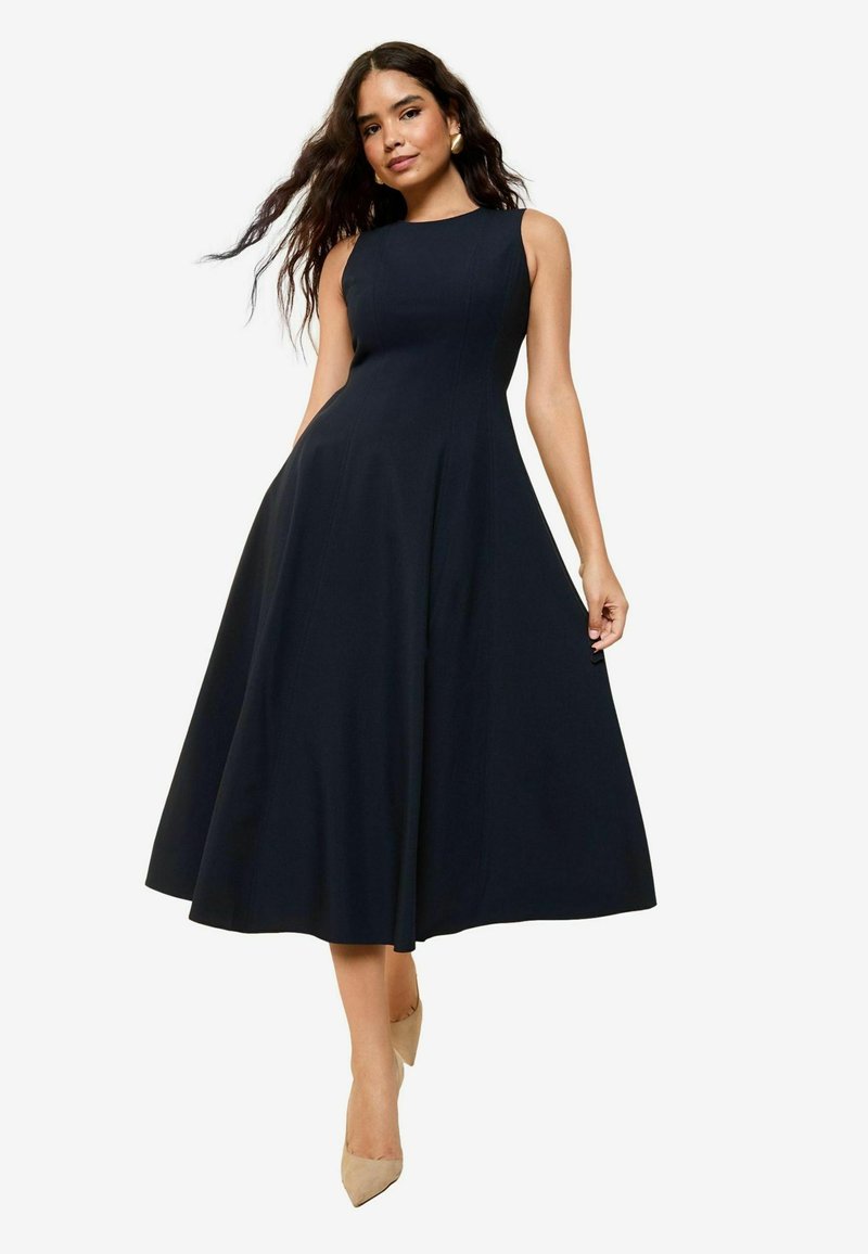 Navy blue sleeveless dress with a fitted bodice and flared skirt, made of smooth fabric. Hemline reaches mid-calf. Footwear is beige.