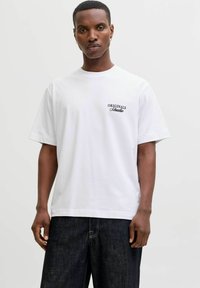 Λευκό βαμβακερό μπλουζάκι με στρογγυλή λαιμόκοψη, κοντά μανίκια και λογότυπο "ORIGINALS" σε μαύρο χρώμα στην αριστερή πλευρά του στήθους. Φορεμένο με σκούρα τζιν παντελόνια.