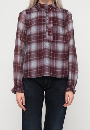 Kobieta ubrana w przezroczystą bluzkę w bordowo-białą kratę z długim rękawem, ozdobioną falbanami przy kołnierzyku i mankietach, zestawioną z ciemnoniebieskimi dżinsami.