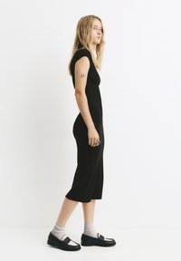 Musta bodycon-mekko lyhyillä hihoilla, polvipituisella helmalla, yhdistettynä mustiin loafereihin vaaleiden sukkien päälle. Malli seisoo profiilissa valkoista taustaa vasten.