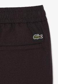Pantalon à taille élastique noire avec une poche passepoilée à l'arrière et un petit logo crocodile vert brodé près de la poche.