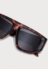 Schildpatt-Sonnenbrille mit einem schwarz-orangefarbenen Muster, rechteckiger Form, dunklen Gläsern und dicken, glatt strukturierten Rahmen.