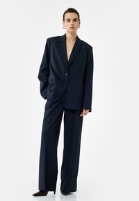 Marineblauw gestreept pak met een lange blazer en wijde broekspijpen. De stof is gestructureerd, met twee knopen en een enkele borstzak.