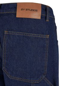 Jean en denim bleu foncé avec une étiquette en cuir portant la marque « 2Y STUDIOS », des coutures oranges et une poche arrière unique.