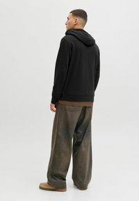 Homme portant un sweat à capuche noir, un pantalon marron surdimensionné et des chaussures beiges, debout légèrement de profil devant un fond clair uni.