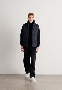 Gilet imbottito senza maniche scuro sopra un dolcevita nero, abbinato a pantaloni neri larghi e sneaker bianche; tessuto liscio e design minimale.