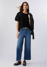 Črna majica z naborki na rokavih in zakovicami, v kombinaciji z širokimi kavbojkami in črnimi sandali z okraski. Usnjena jakna držana v eni roki.