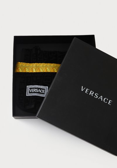 Fekete ajándékdoboz ezüst "VERSACE" felirattal. Egy fekete tárgyat tartalmaz, melyen hangsúlyos sárga mintás szalag és fehér logófolt található.