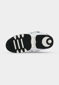 Semelle d'une chaussure de sport blanche et noire montrant un motif de bande de roulement avec des crampons circulaires et rectangulaires et le logo de la marque au centre.