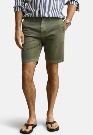 Mann trägt olivgrüne Shorts, gestreiftes Langarmshirt, silberne Armbanduhr und braune Sandalen, steht mit einer Hand in der Tasche.
