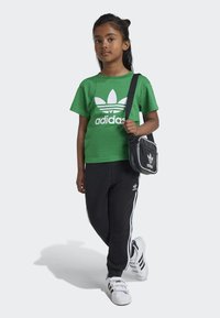 Zelené bavlněné tričko s bílým logem Adidas, černé tepláky s bílými bočními pruhy a černá crossbody taška s logem a lemováním.