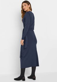 Marineblaues langärmliges Kleid mit Wickeldesign, seitlichem Bindeband und lockerem Schnitt. Getragen mit schwarzen Strumpfhosen und niederen Schuhen. Einfach und elegant.