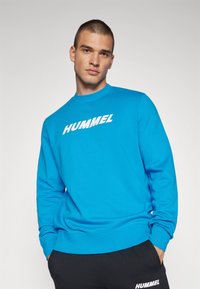 Niebieska bluza z ściągaczami przy mankietach i dekolcie, z dużym białym logo "HUMMEL" na piersiach, w połączeniu z czarnymi spodniami.