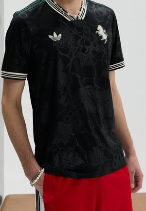 Personne portant un t-shirt Adidas noir avec des motifs floraux foncés, un logo blanc et des bandes sur le col et les manches, associé à un pantalon rouge et des bracelets en perles.