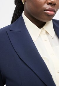 Κοντινό πλάνο ενός ατόμου που φοράει μπλέ σκούρο σακάκι πάνω από λευκό πουκάμισο με κουμπιά, με χρυσούς κρίκους σκουλαρίκια να φαίνονται σε λευκό φόντο.