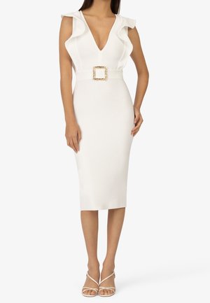 Mujer con un vestido midi blanco ajustado con mangas cortas con volantes, escote en V profundo, cinturón con hebilla dorada y tacones blancos con tiras, sobre un fondo blanco.