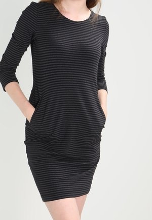 Kobieta ubrana w dopasowaną sukienkę w poziome paski czarno-antracytowe z rękawami do połowy przedramienia i rękami w bocznych kieszeniach.