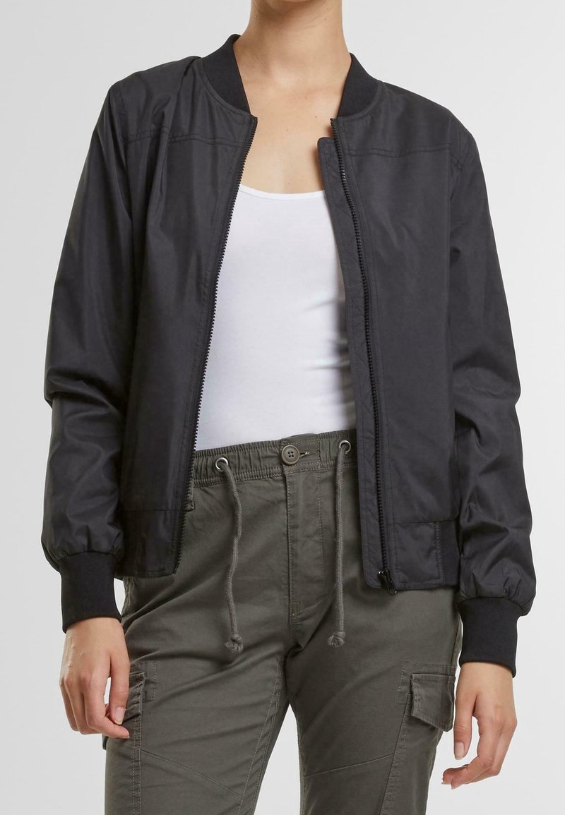 Personne portant une veste bomber noire zippée sur un débardeur blanc et un pantalon cargo vert olive avec des cordons.