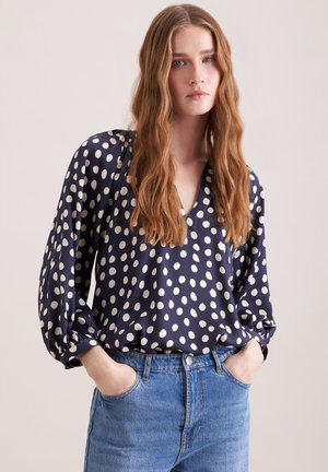 Femme aux longs cheveux ondulés portant un chemisier bleu marine à pois blancs et un jean bleu, les mains dans les poches, sur un fond uni.