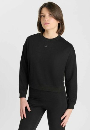 Giovane donna con capelli corti e castani che indossa un completo totalmente nero, che include una maglietta a maniche lunghe e pantaloni aderenti, in piedi davanti a uno sfondo uniforme.