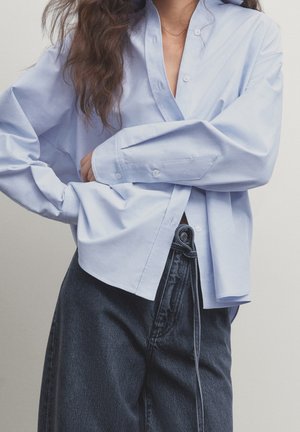 Woman with long wavy hair wears a loose light blue button-up shirt and high-waisted dark denim jeans with a tied belt.