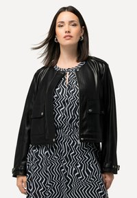 Schwarze Lederimitat-Cropped-Jacke mit einem Frontreißverschluss, zwei Brusttaschen und Druckknopf-Akzenten. Tragen über einem gemusterten schwarz-weißen Kleid.