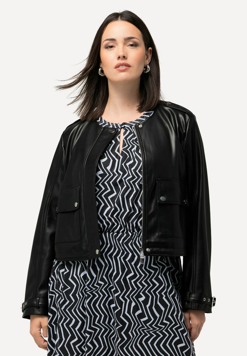 Schwarze Lederimitat-Cropped-Jacke mit einem Frontreißverschluss, zwei Brusttaschen und Druckknopf-Akzenten. Tragen über einem gemusterten schwarz-weißen Kleid.
