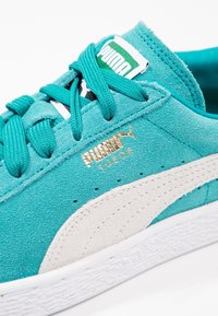 Turkusowe zamszowe sneakersy z białymi zamszowymi akcentami, płaskimi turkusowymi sznurowadłami i złotym logo po boku, z teksturowaną białą gumową podeszwą.