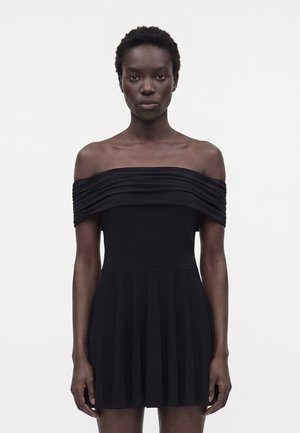 Mujer con el cabello corto, vestida con un vestido negro plisado sin hombros, de pie frente a un fondo claro y liso, mirando al frente.