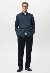 Camisa azul marino de botones con tela texturizada, combinada con pantalones oscuros y zapatos negros. La camisa presenta un cuello puntiagudo y mangas largas.