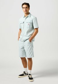 Camisa de manga corta azul claro con rayas verticales y dos bolsillos frontales, combinada con pantalones cortos a juego. El modelo lleva zapatillas negras y calcetines blancos.