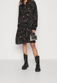 Czarna sukienka z kolorowymi abstrakcyjnymi wzorami, długimi rękawami, zapięciem na guziki i rozkloszowanym dołem. Połączona z czarnymi butami na grubym obcasie i srebrną torebką.