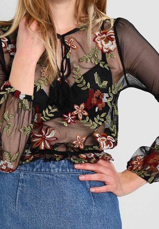 Blusa nera trasparente con ricami floreali colorati, maniche lunghe con polsini arricciati e un dettaglio a fiocco sullo scollo.