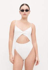 Maillot de bain blanc une pièce avec un devant noué, des côtés découpés, des fines bretelles et un tissu texturé. Accompagné de lunettes de soleil noires.