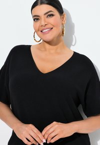 Černý pletený top s výstřihem do V a krátkými rukávy. Má strukturovanou látku a volný střih. Modelka má ruce sepnuté před sebou.