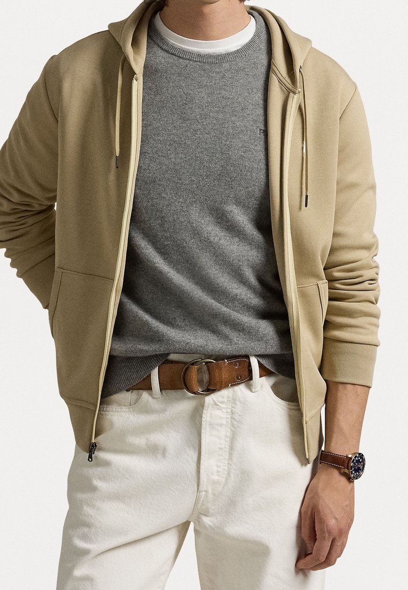 Homme portant un sweat à capuche beige zippé sur un pull gris, un pantalon blanc avec une ceinture marron et une montre-bracelet en cuir marron.