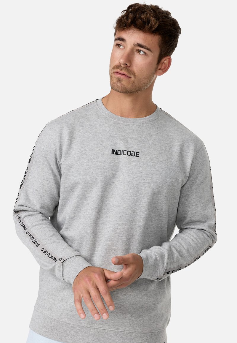 Sweatshirt gris en coton, avec des manches longues. Logo noir "INDICODE" au niveau de la poitrine et sur les coutures latérales. Poignets et ourlet côtelés.