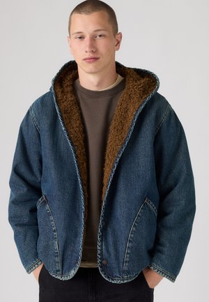 Giovane uomo che indossa una giacca di jeans blu con fodera in pile marrone sopra una felpa marrone, in piedi con le mani in tasca davanti a uno sfondo semplice.