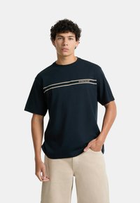 T-shirt di cotone blu navy con maniche corte, caratterizzato da una striscia orizzontale beige e il testo "GUESS JEANS" sul petto.
