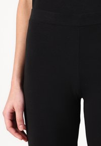 Leggings pretos com uma textura suave e um cós elástico, mostrando parte de uma mão descansando levemente no material.