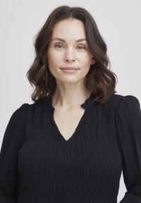 Schwarze Bluse mit einem strukturierten, gerafften Design, einem V-Ausschnitt und einem Rüschenausschnitt. Lange Ärmel mit subtilen elatischen Bündchen.