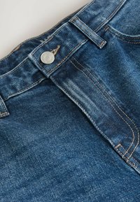 Denim teksad, millel on klassikaline sinine pesu, nähtavad õmblused, hõbedane nupuga kinnitamine ja traditsiooniline viie taskuga disain.