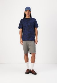 Cămașă bleu închis cu mânecă scurtă, cu un model de palmieri, asortată cu pantaloni șort gri, șosete albe și mocasini maro. Se poartă o șapcă albastră.
