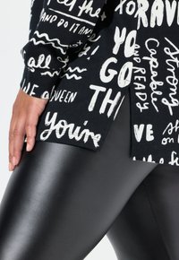 Sudadera negra de manga larga con patrones de texto en blanco, combinada con leggings negros brillantes. La sudadera cuenta con una abertura lateral y una textura de tela suave.