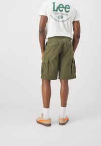 Bermudas cargo de color verde oliva con múltiples bolsillos laterales y un corte relajado, combinadas con una camiseta blanca con un logo verde, calcetas blancas y zapatillas beige.