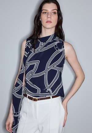 Jeune femme portant un haut sans manches bleu marine avec un imprimé chaîne blanc, rentré dans un pantalon blanc taille haute avec une ceinture marron, la main dans la poche.