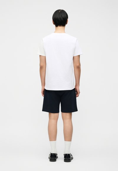 Άτομο στέκεται με την πλάτη γυρισμένη, φοράει λευκό κοντομάνικο πουκάμισο, μαύρο σορτς, λευκές κάλτσες και μαύρα παπούτσια πάνω σε ένα απλό ανοιχτό φόντο.