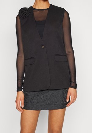 Femme portant un blazer noir sans manches avec un détail fleur, un top noir transparent à manches longues et une mini-jupe noire texturée.