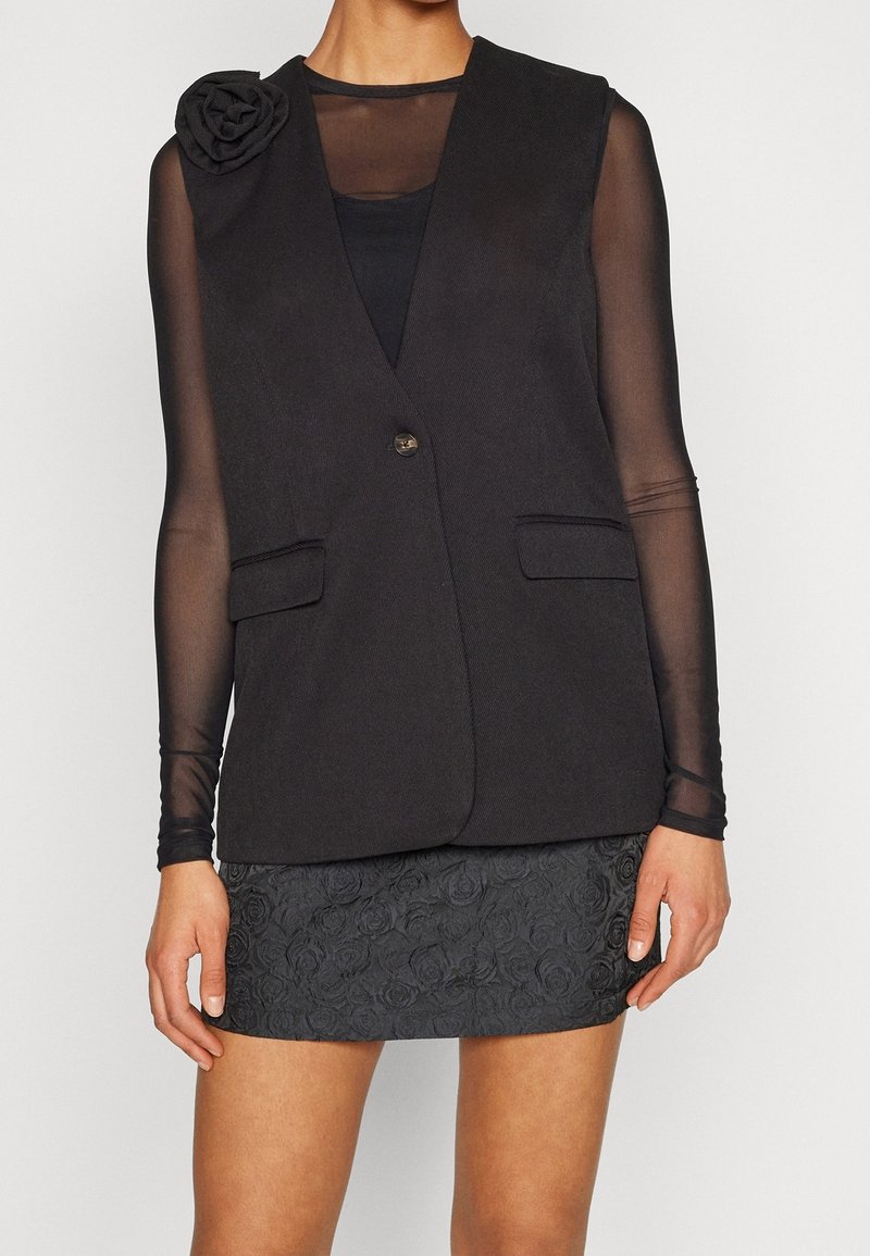 Femme portant un blazer noir sans manches avec un détail fleur, un top noir transparent à manches longues et une mini-jupe noire texturée.